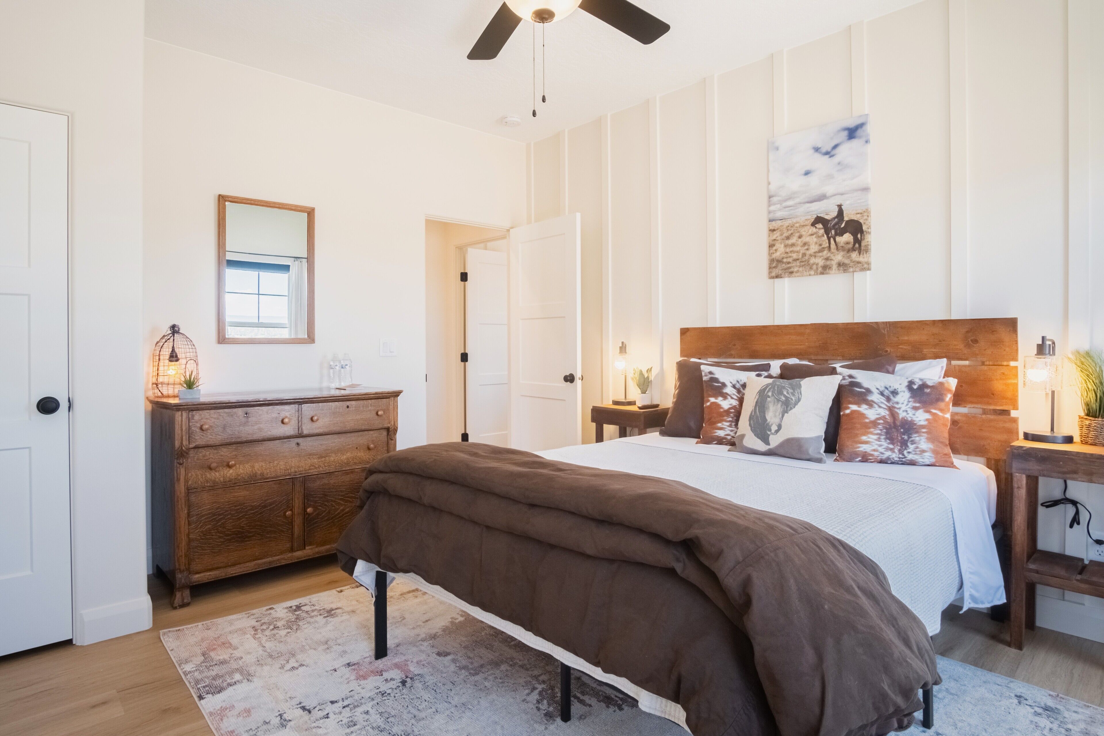 A bedroom with a large bed, wooden headboard, a ceiling fan, a nightstand with a lamp, a dresser, and a painting on the wall.