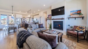 A spacious living area with a fireplace, a flat-screen TV, and a dining area with a kitchen in the background. - Kanab Getaway Red Mesa Ranch, Near Zion, Amazing Red Rock & Starry Night Views! (Kanab ( Will Show as Canyon City on Some Apps))