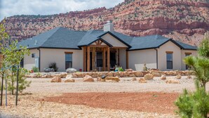 A two-story house with a stone foundation and a dark roof, surrounded by a landscaped area with rocks and sparse vegetation. - Kanab Getaway Red Mesa Ranch, Near Zion, Amazing Red Rock & Starry Night Views! (Kanab ( Will Show as Canyon City on Some Apps))