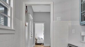 Ein schmaler Flur mit weiß gefliesten Wänden und Holzboden, der zu einem Schlafzimmer mit Bett und Fenster führt.