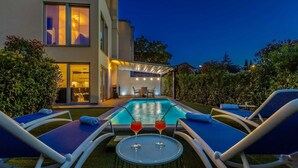 A modern outdoor pool area with lounge chairs and a table with drinks, surrounded by greenery and a building with large windows. - Modern villa MODERNA with heated pool, hot tub and sauna (Malinska)