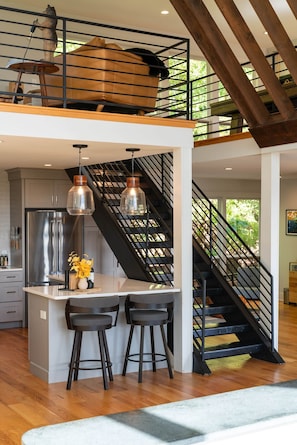 A modern kitchen with a central island, two bar stools, a stainless steel refrigerator, and pendant lighting. - Greenwood Vista (Greenwood)