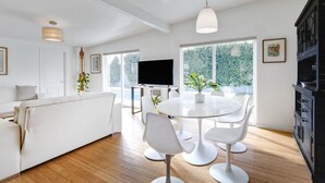 A modern living room with a white sofa, a dining table with chairs, a flat-screen TV, and large windows offering a view of greenery. - ICONIC MID-CENTURY MODERN  “HAVER HOME” (Scottsdale)
