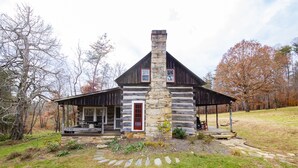 Ein rustikales Blockhaus mit einem Steinkamin, einer rotem Tür und einer Veranda mit Sitzgelegenheit.