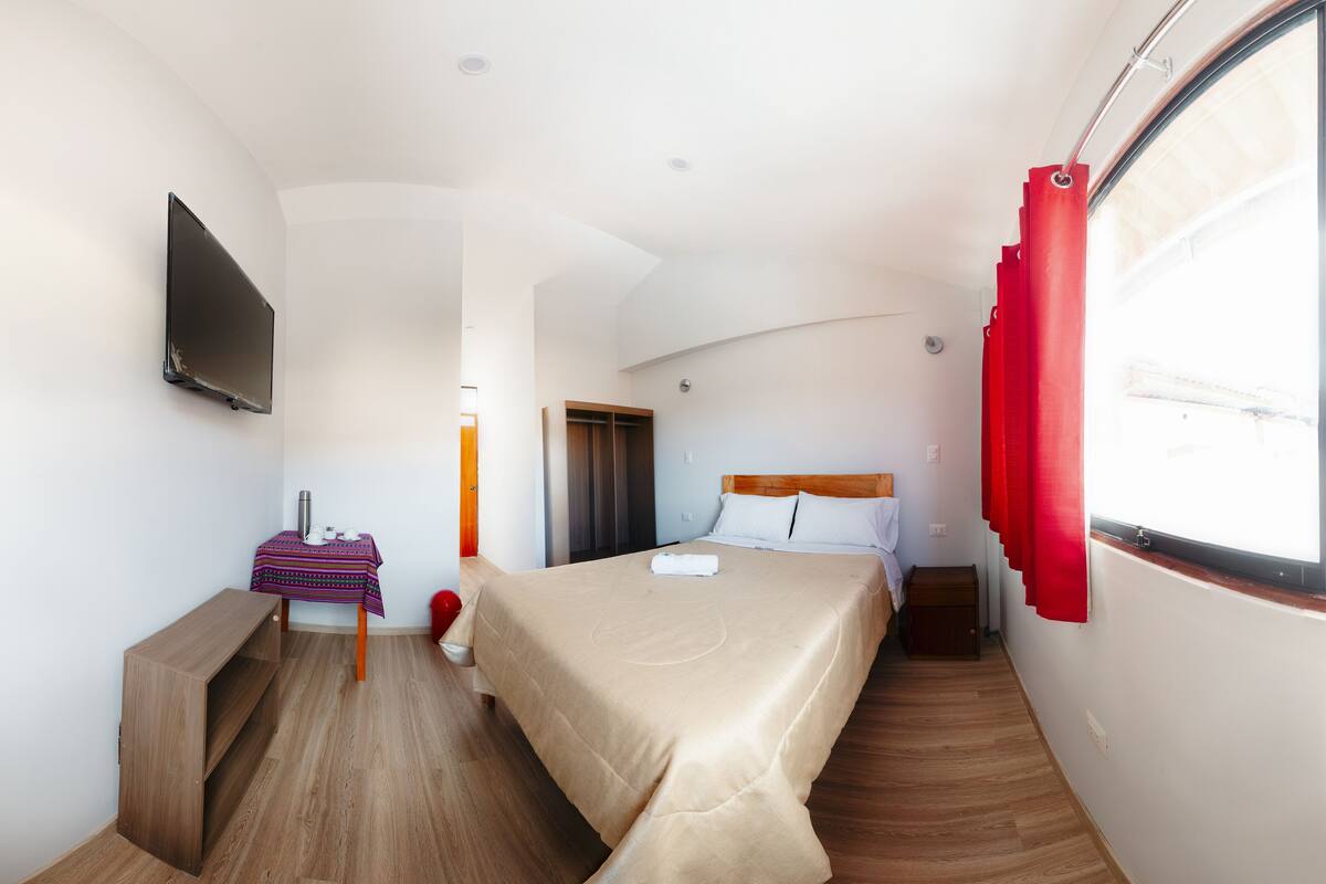 Habitación de hotel con cama, televisor montado en la pared, una mesita con mantel y un banco.