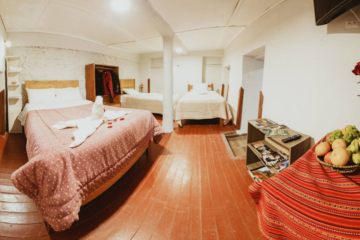 Habitación de hotel con dos camas, piso de madera, pared de piedra y una pequeña mesa con una cesta de frutas.