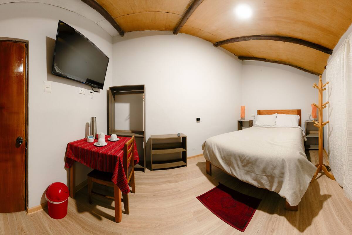 Un dormitorio con cama, televisor montado en la pared, techo de madera y alfombra roja.
