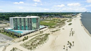 A modern hotel with a glass facade, located adjacent to a beach with a sandy shoreline and sparse palm trees. - Coveted Gulf-front condo on pool deck, directly on beach, no highway to cross! (Biloxi)