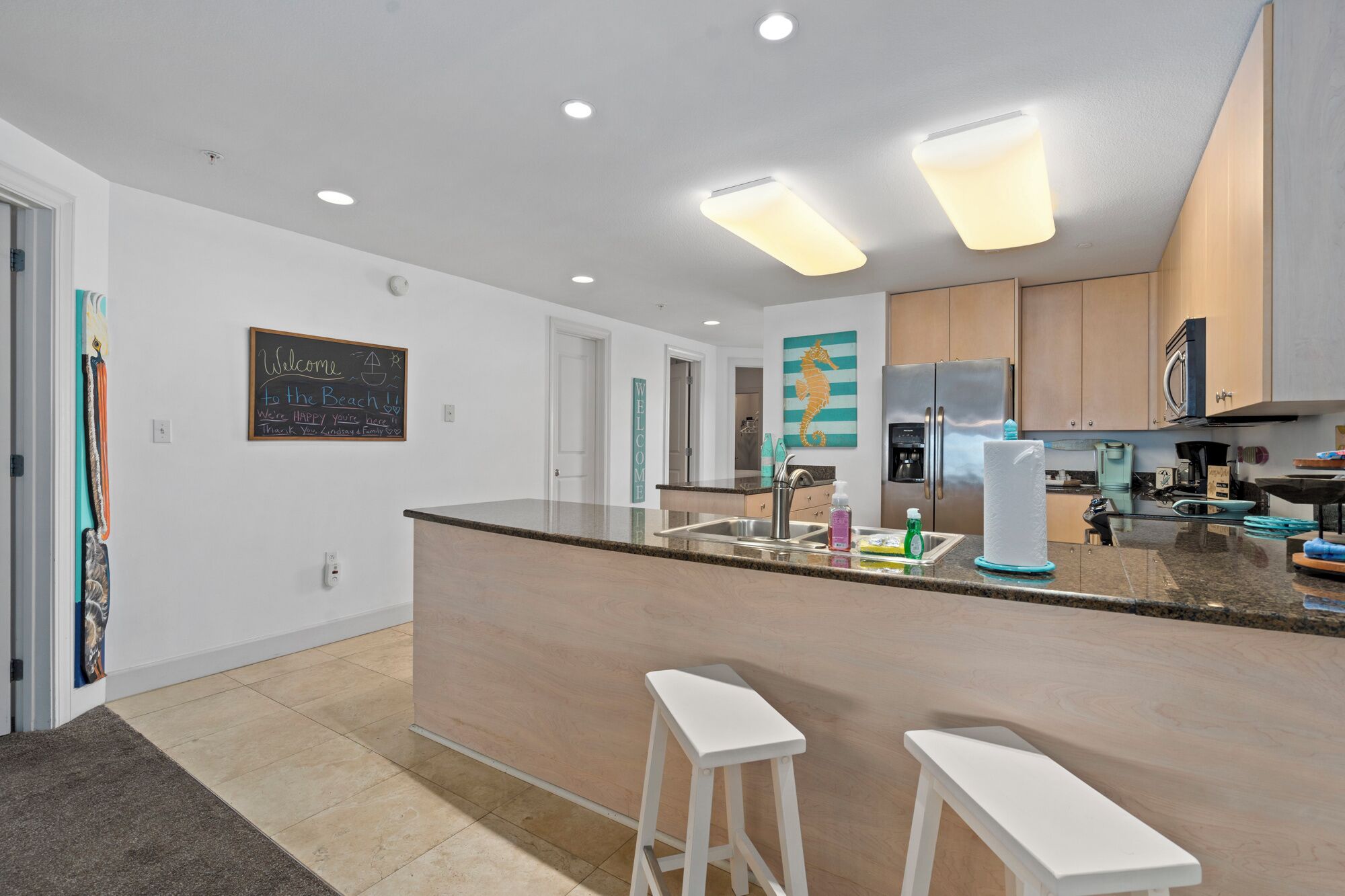 A modern kitchen with a central island, stainless steel appliances, and a chalkboard with a welcome message.
