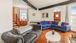 A living room with a grey sofa, a blue sectional sofa, a white coffee table, and a wooden floor. There is a wooden staircase on the left, a window on the right, and a door with a mirror above it. - Victorian style home, where shabby chic meets the beach (Torquay)