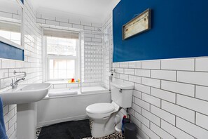 Une salle de bain avec des carreaux de céramique blancs rectangulaires, un mur d’accent bleu, une fenêtre, un lavabo, des toilettes et une douche.