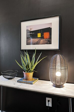 Ein gerahmtes Foto an der Wand, ein Topfpflanze, ein Buch, ein Drahtkorb und eine strukturierte Lampe auf einer Marmorfläche.