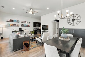 A modern living room with a grey sofa, a dining table with chairs, a kitchen area with a microwave, and a large wall clock.