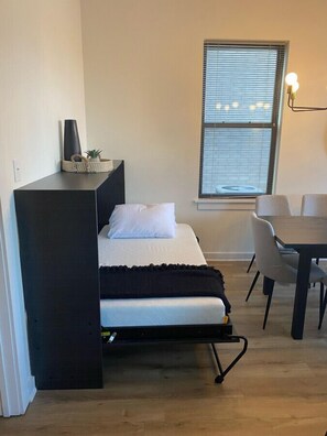 Ein kleines Zimmer mit einem Bett, einem Tisch und einem Fenster mit Jalousien.