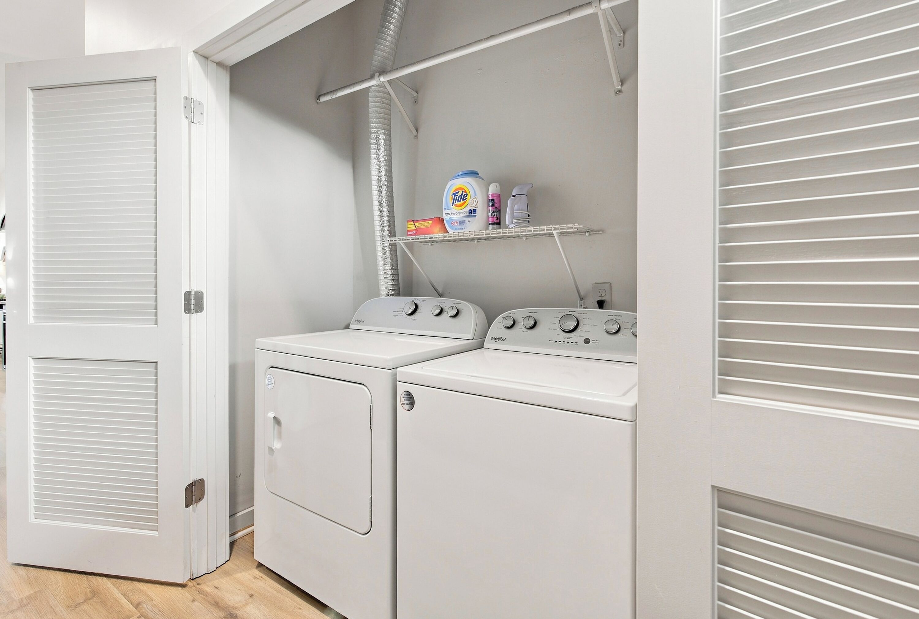A laundry room with two Whirlpool washers and dryers, a shelf with cleaning products, and a vent system.