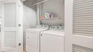 A laundry room with two Whirlpool washers and dryers, a shelf with cleaning products, and a vent system.