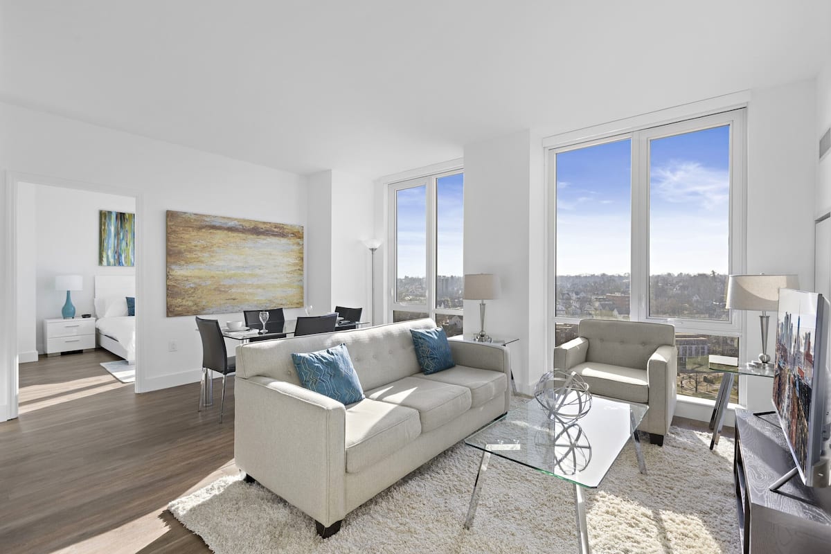 A modern living room with a sofa, armchairs, a glass coffee table, and a city view through large windows.