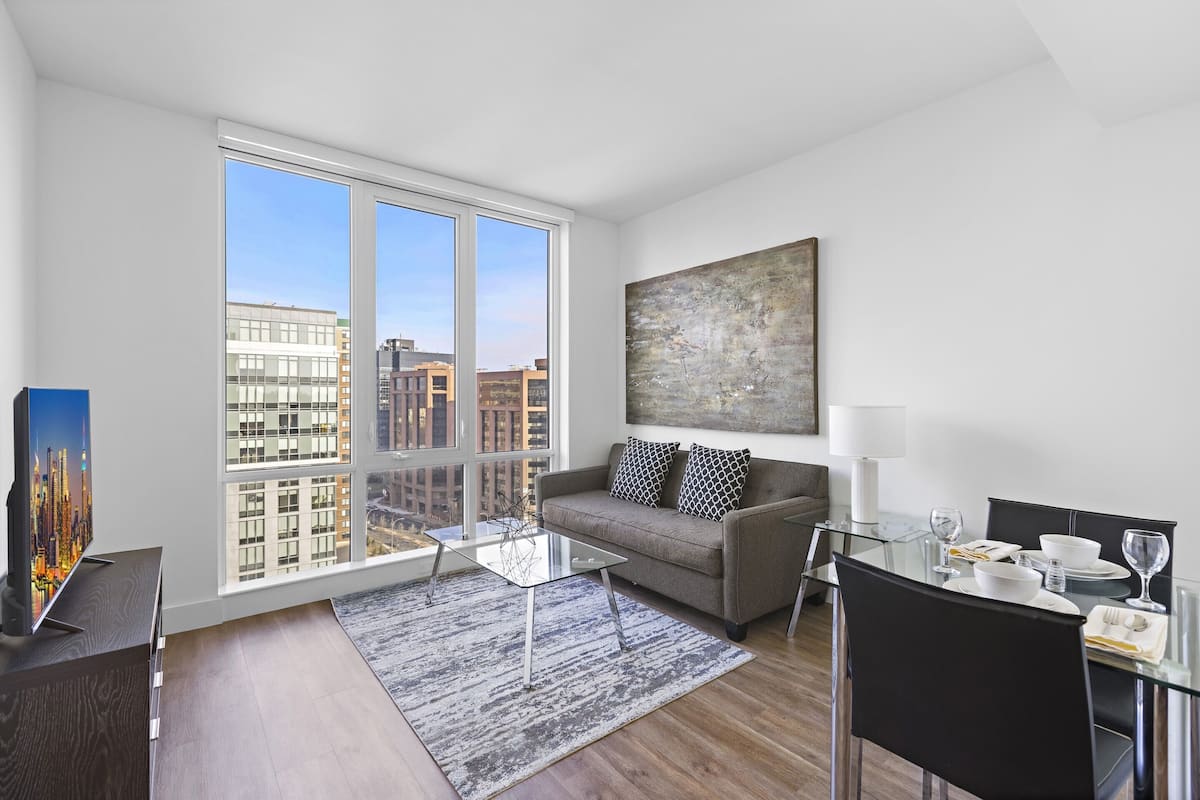 A modern living room with a large window, a sofa, a glass coffee table, and a dining area with a table and chairs.