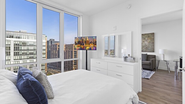 Un dormitorio moderno con una cama grande, televisión, cómoda y vistas a un paisaje urbano a través de amplios ventanales.
