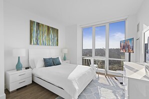 A modern bedroom with a large bed, white nightstands, a desk, and a large window offering a city view. - Global Luxury Suites White Plains (White Plains)
