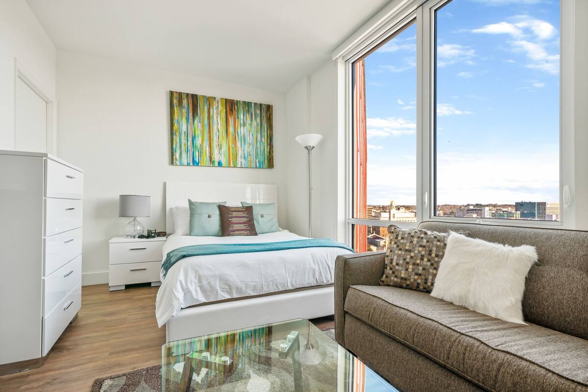 A modern bedroom with a large window, a bed, a sofa, and a desk with a lamp.