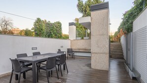 Une terrasse extérieure avec une table et des chaises, un pilier en pierre et un escalier menant à une zone abritée.