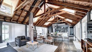 Ein geräumiger Wohnbereich mit hoher Decke, freiliegenden Holzbalken, einem Kamin und einer modernen Küche im Hintergrund.