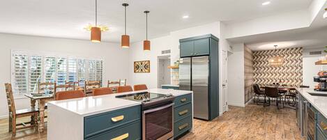 A modern kitchen with a central island, built-in appliances, pendant lighting, and a dining area with wooden chairs.