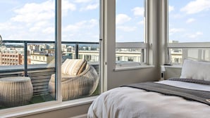 A bedroom with large windows, a bed, a wicker chair, and a view of the cityscape. - Luxurious, Ocean View 2 Story Penthouse - Downtown - Secure Parking (Victoria)