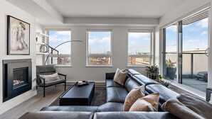 A modern living room with a sofa, a coffee table, and a fireplace. There are large windows offering a view of the city. - Luxurious, Ocean View 2 Story Penthouse - Downtown - Secure Parking (Victoria)