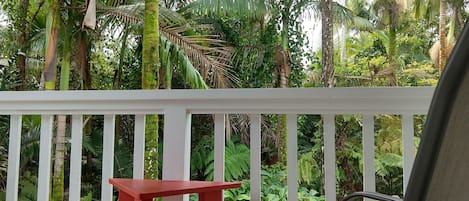 Un balcón blanco con una mesa roja y dos sillas, rodeado de exuberante vegetación tropical.