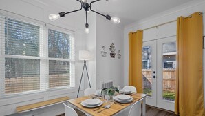 A dining area with a wooden table set for four, white chairs, a floor lamp, and a view through a door with yellow curtains.