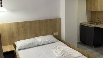 A bedroom with a wooden headboard, a bed with white bedding, a cat-themed wall decal, a bedside table, and a small kitchenette with a sink and cabinets.