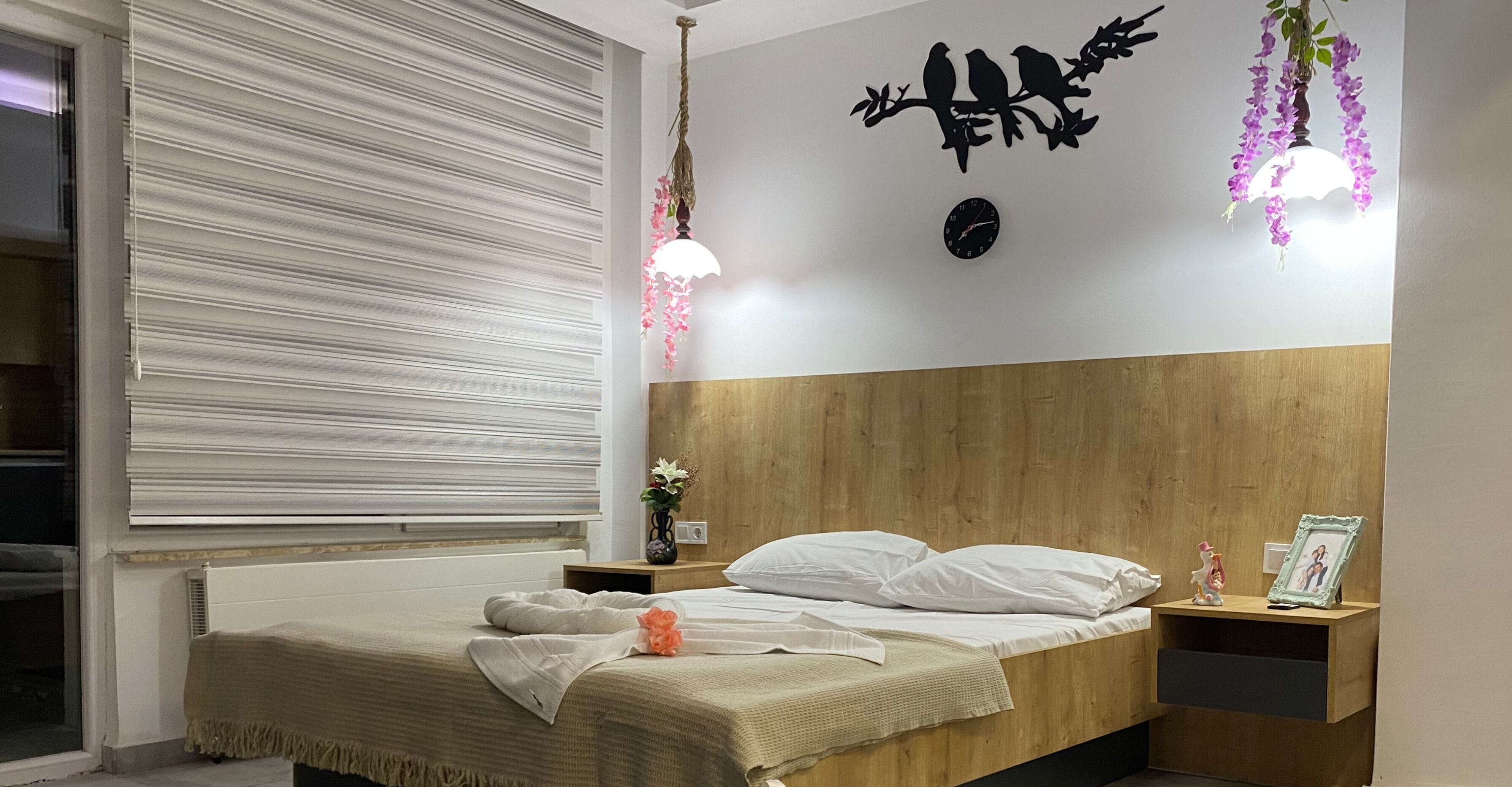 A bedroom with a bed, a nightstand, a wall clock, and a window with blinds.