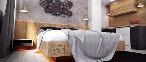 A modern bedroom with a wooden headboard, a bed with a grey blanket, a wall-mounted gear decoration, and a kitchenette in the background.