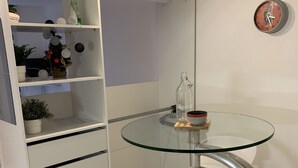 A compact room with a glass table and stools, a white shelf unit with decorative items, and a wall clock.