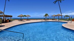 A swimming pool with a view of the ocean, surrounded by palm trees and thatched umbrellas. - Spacious OceanFront 2BD w Pool AC-Royal Kahana 701 (Lahaina)