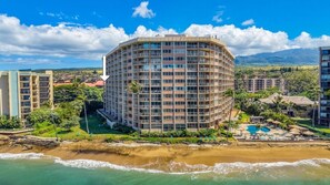 A multi-story hotel with a curved facade, located adjacent to a beach with a pool and lounge area.