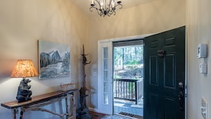 A wooden console table with a bear figurine and a lamp, a painting of mountains, a tree sculpture, and a dark green door leading to a balcony.