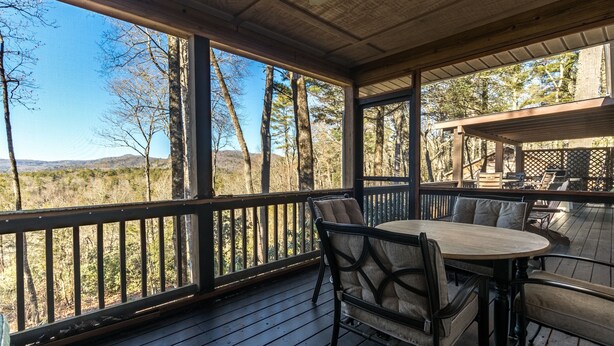 A covered outdoor seating area with a table and chairs, overlooking a forested landscape.