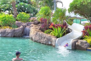 A pool area with a water slide, rocks, and tropical plants.