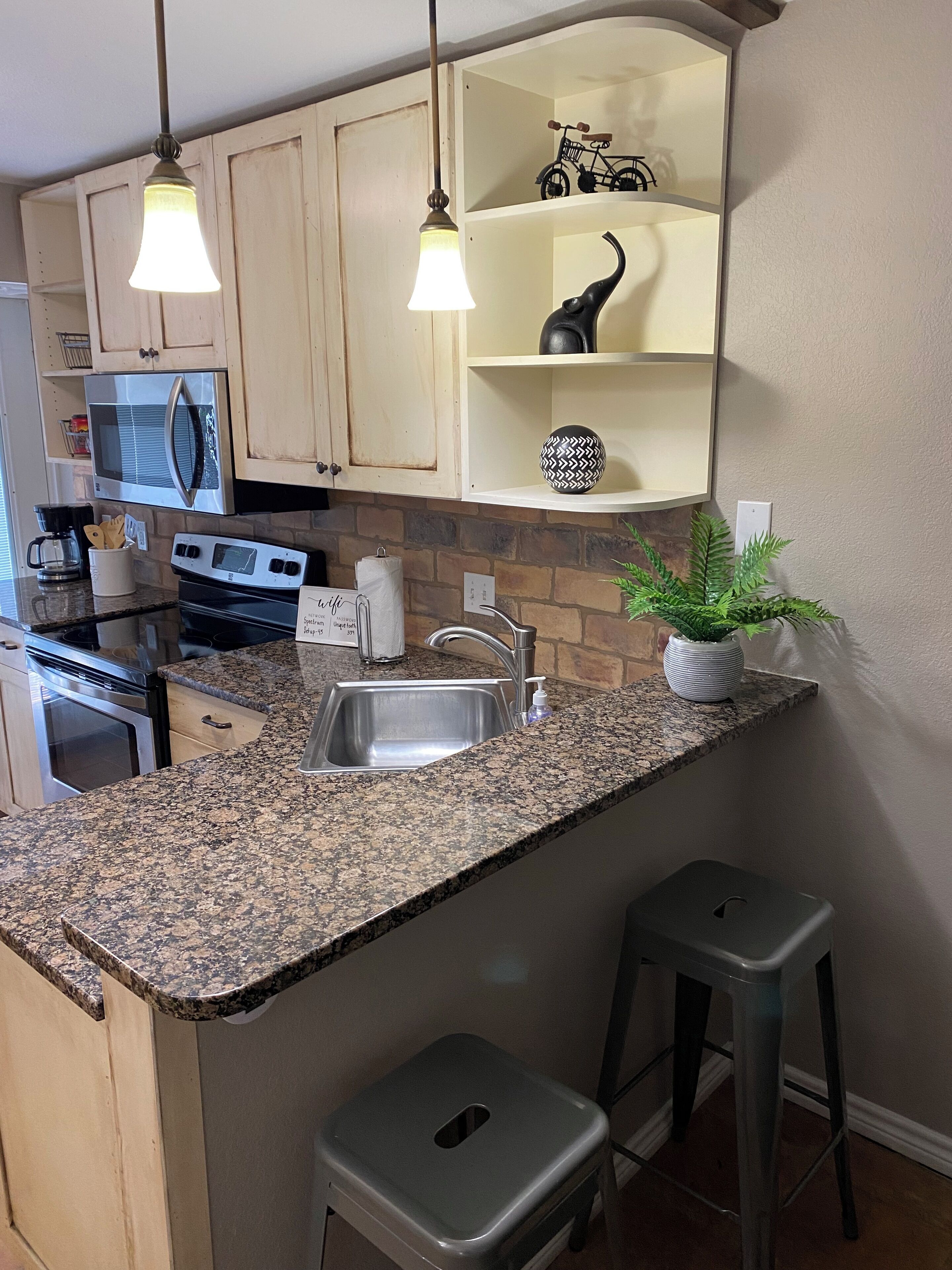 A kitchen with granite countertops, stainless steel appliances, and a built-in oven. There are two bar stools and a potted plant on the counter.