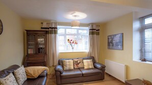 A living room with a brown leather sofa, a wooden cabinet, a radiator, and a window with curtains. - 2 bedroom accommodation in Oswestry (Oswestry)