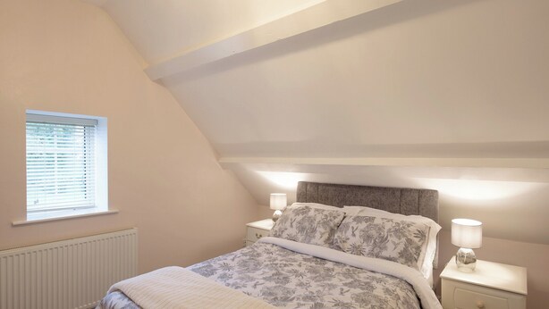 A bedroom with a sloped ceiling, a window with blinds, a bed with floral bedding, two bedside tables with lamps, and a radiator. - 2 bedroom accommodation in Oswestry (Oswestry)
