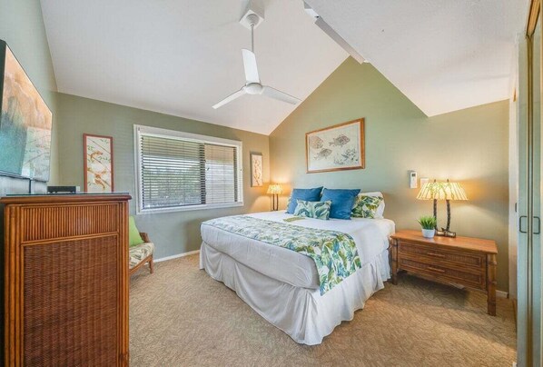 A bedroom with a bed, a ceiling fan, a window with blinds, a bedside table with a lamp, and a wall-mounted TV.