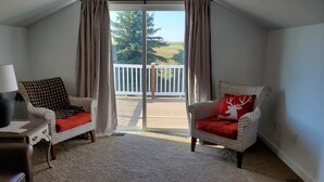 A room with two wicker chairs, one with a red cushion and the other with a checkered cushion, a small table with a lamp, and a view of a deck with a white railing and greenery outside. - Historic ranch house has beautiful mountain and valley views. Weekly discounts. (Manhattan)