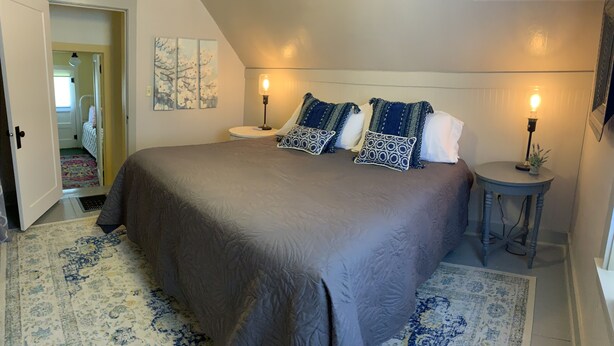 Une chambre avec un grand lit, deux tables de chevet avec des lampes, une porte donnant sur une autre pièce et un tapis à motifs.