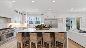 A modern kitchen with white cabinetry, a central island with bar stools, stainless steel appliances, and a view to the outside. - Luxurious 4-BR Home w/ Private Pool & Beach Access – Golf Cart & Bikes Included (Santa Rosa Beach)