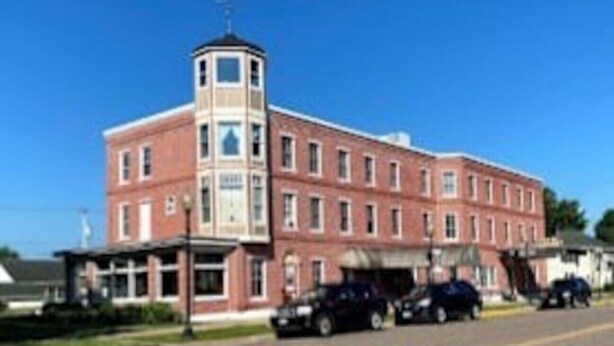A multi-story brick building with a central tower, a covered entrance, and several parked cars in front. - AH #11 · AH #11 Queen Suite w/Tub/Shower (Wabasha)