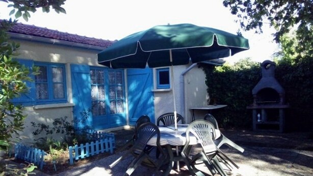 Een terras met een tafel en stoelen onder een groene parasol, een kleine buitenoven en een blauw huis met een witte deur.
